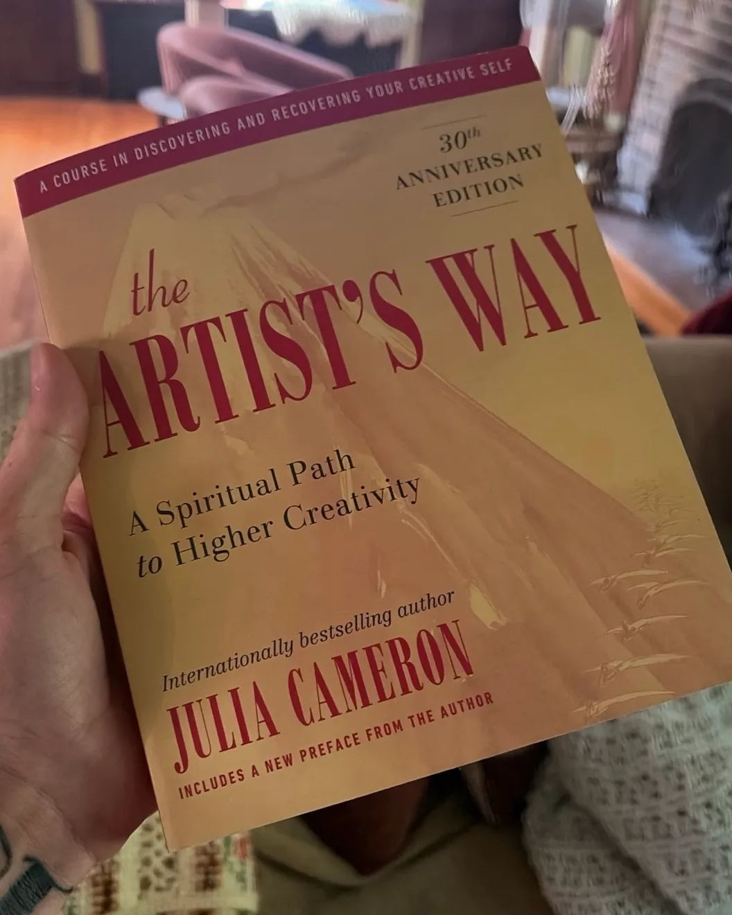 Joe's hand holding a copy of The Artist's Way by Julia Cameron, 30th anniversary edition, in his living room