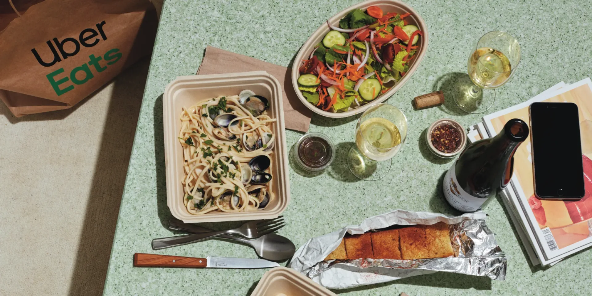 Uber Eats takeout spread: clam linguine, a salad, garlic bread and white wine on a green table