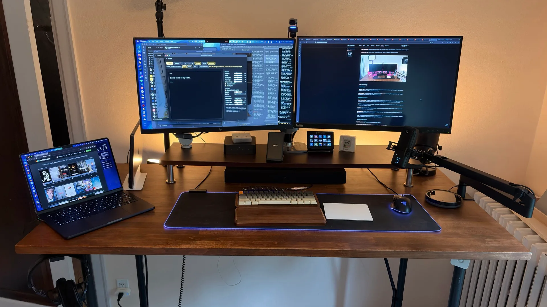 Close-up front view of Joe's desk: two monitors on a riser with warm LED backlighting, a Stream Deck, an Aranet4 CO2 monitor, a mechanical keyboard with a wood wrist rest, and an RGB mousepad