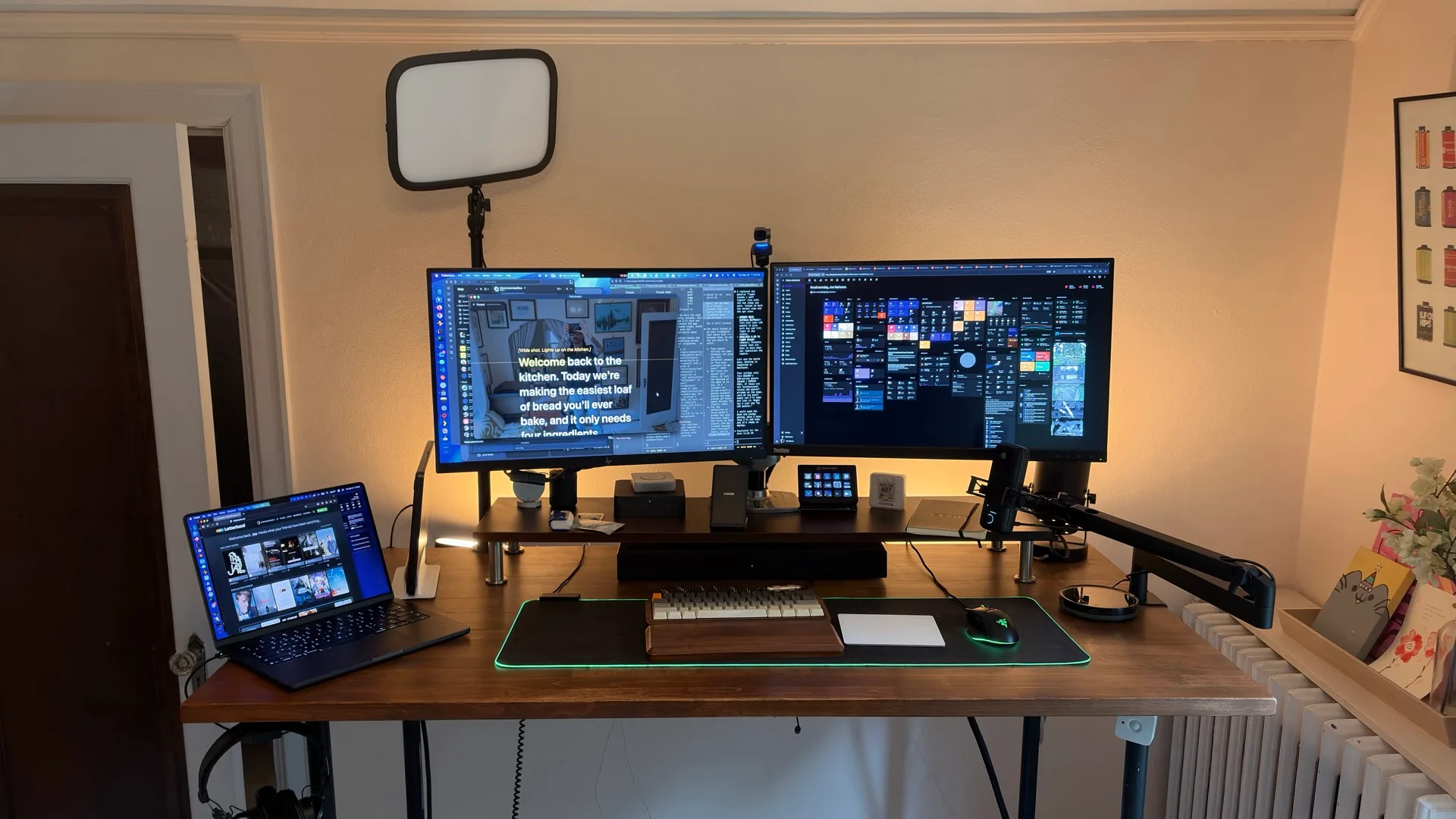 Joe Karlsson's standing desk with two monitors, a laptop on a stand, a mic on a boom arm, a key light above the monitors, and warm LED backlighting