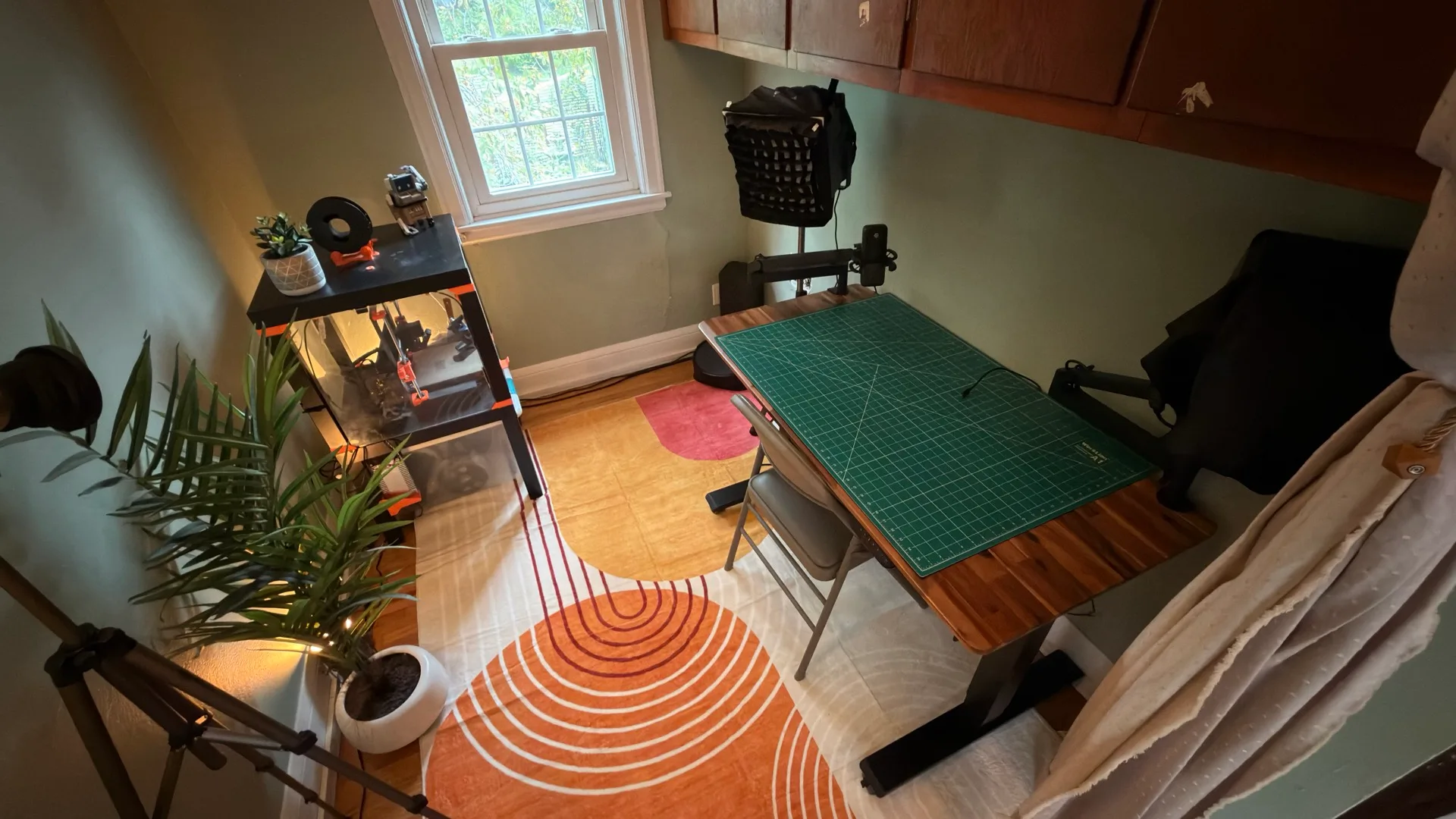 Overhead view of Joe's recording studio: an acacia standing desk with a green cutting mat, a softbox light, a mic and camera on boom arms, a chair, a shelf with a 3D printer, and a palm plant